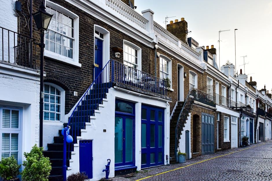A row of terraced houses on a cobbled street, featuring dark brick facades and white window frames. One property has a blue front door and a small balcony with a matching blue railing, accessed by a staircase that extends from the street level. The front area includes various packaging materials such as cardboard boxes and plastic wrap, along with furniture like a wooden table and chairs visible through large windows. A man is seen lifting a cardboard box, possibly during the packing or loading process of home relocation. Nearby, a van is partially visible, parked close to the curb, ready for furniture transport. The environment is well-lit with natural daylight, and the scene captures the logistical aspects of packing and moving, with the company Man and Van Notting Hill providing removal services as part of a house move in the area.