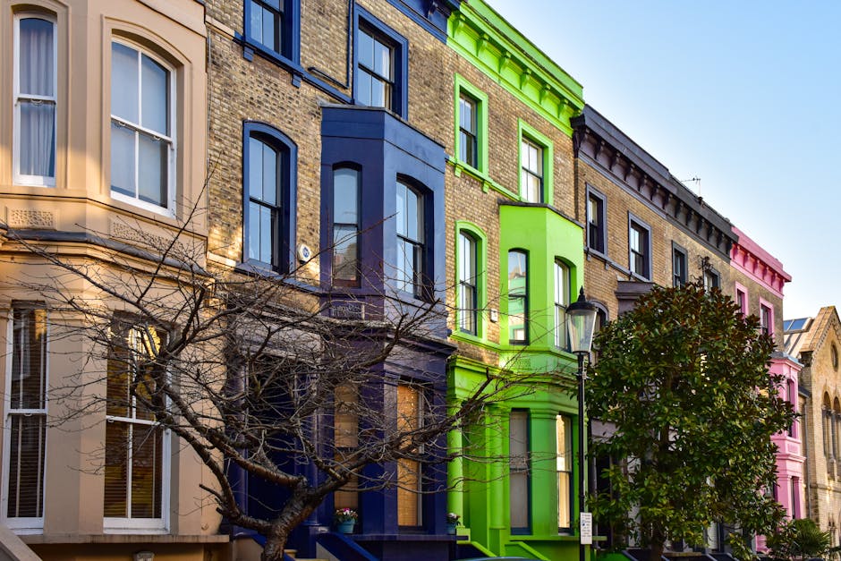 A row of colourful Victorian-style terraced houses in Notting Hill, London, with facades painted in cream, blue, green, and pink, featuring sash windows and decorative cornices. A leafless tree with branches extending across the lower part of the scene partially obscures the buildings. A black street lamp is positioned in front of the green house, and the bright daylight sky enhances the vibrant appearance of the buildings. This image depicts a typical residential street often used for home relocations, with the facade showing the exterior environment where moving activities, such as furniture transport or packing, might take place nearby, consistent with house removals services provided by companies like Man and Van Notting Hill.