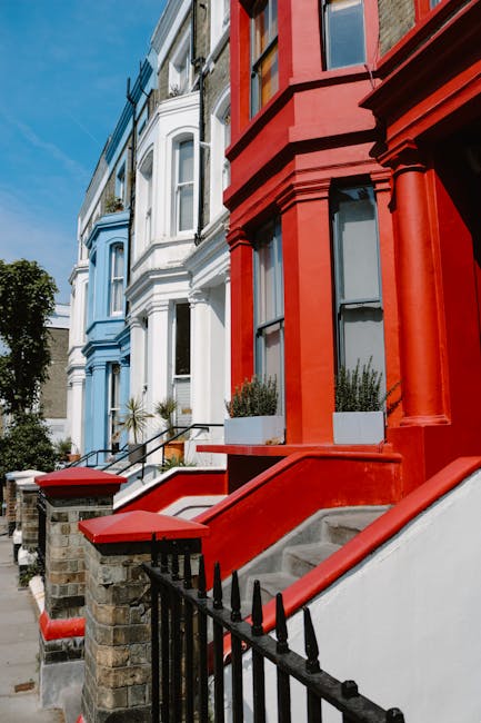 A row of colorful Victorian terraced houses on a urban street, featuring detailed architectural elements and large sash windows. The house in the foreground is painted bright red with white accents around the windows and a small front garden area with rectangular planters containing greenery. Concrete stairs with a metal railing lead up to the red front door, and additional houses further down the street are painted in shades of blue and white. The pavement is visible, with some shadowed areas cast by nearby trees. The scene appears well-lit with natural daylight, capturing the typical residential character of Notting Hill, which is often associated with home relocations or furniture transport services provided by companies like Man and Van Notting Hill. This setting illustrates a typical London street where careful packing, loading, and moving logistics are necessary during house removals and furniture transport jobs.