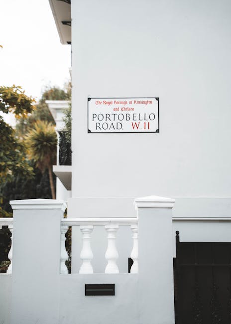 A close-up view of a white residential building facade showing a street sign for Portobello Road, located on the corner of a property in Notting Hill, Kensington & Chelsea. The sign indicates Load rules apply and features the Royal Borough of Kensington and Chelsea emblem. In the foreground, a white fence with decorative balusters and a black mailbox are visible, suggesting a front garden or entrance area. The building's exterior wall is smooth and painted white, with natural lighting highlighting the clean surfaces. Part of a balcony or overhang can be seen on the upper left corner. The scene is set in an urban environment with some greenery, including trees with autumnal foliage, visible in the background. This image relates to property addresses and moving logistics in the Notting Hill area, where home relocation and furniture transport often involve navigating local street regulations.