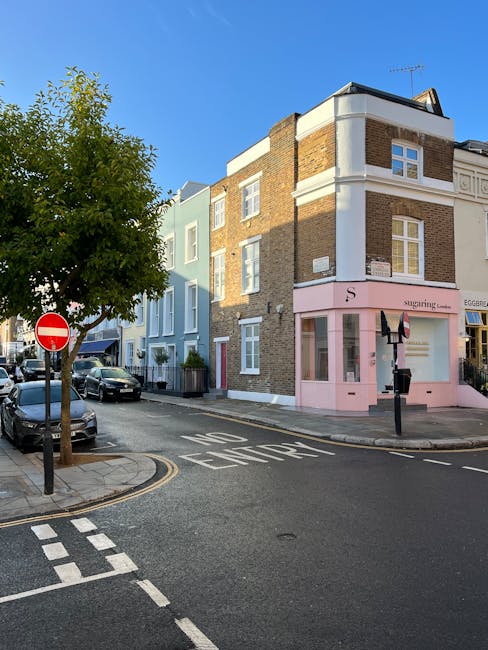 A street scene in Notting Hill featuring a corner building with a pink storefront and upper residential windows, adjacent to a light blue building. The road is marked with 'NO ENTRY' and is lined with parked cars along the pavement. A large leafy tree is situated on the left side of the image, casting shadows on the street. In the foreground, a black bollard and a ‘no entry’ traffic sign are visible, with the pavement showing designated loading zones and road markings. The clear blue sky provides natural lighting, indicating daytime. This scene depicts typical urban street layout relevant to house relocations and furniture transport logistics, as shown in the context of Kensington & Chelsea parking and loading regulations, with elements consistent with moving or packing activities supported by Man and Van Notting Hill.