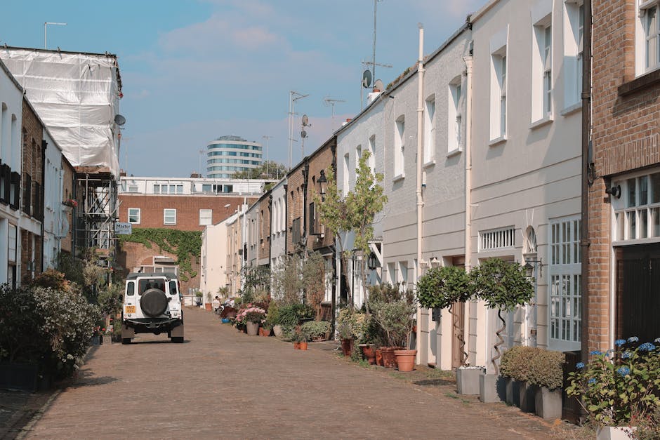 A close-up view of a white residential building facade showing a street sign for Portobello Road, located on the corner of a property in Notting Hill, Kensington & Chelsea. The sign indicates Load rules apply and features the Royal Borough of Kensington and Chelsea emblem. In the foreground, a white fence with decorative balusters and a black mailbox are visible, suggesting a front garden or entrance area. The building's exterior wall is smooth and painted white, with natural lighting highlighting the clean surfaces. Part of a balcony or overhang can be seen on the upper left corner. The scene is set in an urban environment with some greenery, including trees with autumnal foliage, visible in the background. This image relates to property addresses and moving logistics in the Notting Hill area, where home relocation and furniture transport often involve navigating local street regulations.