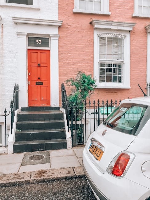 Exterior view of a residential property at 53 Ladbroke Grove with a bright red front door accessed by black stairs with white borders, flanked by a black metal railing. A small green bush is situated beside the stairs, near a window with a white frame and decorative trim. A white Fiat 500 is parked on the street in front of the house, partially visible, with a UK license plate. The pavement is grey concrete with a metal drain cover near the curb. The scene suggests an urban area suitable for home relocation and furniture transport, consistent with professional removals services offered by Man and Van Notting Hill.