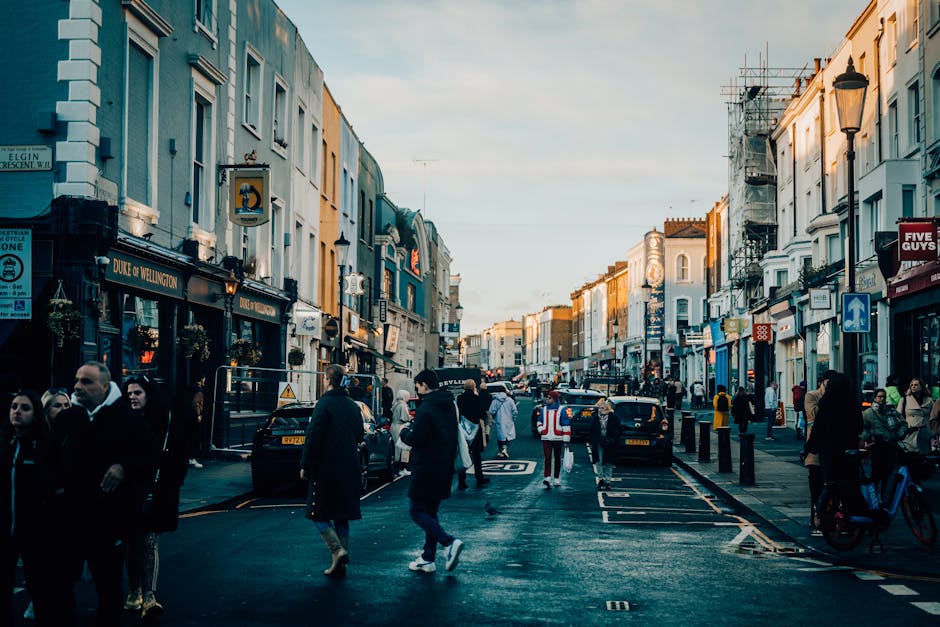 A street scene in Notting Hill featuring a row of colorful terraced houses painted in pastel shades of blue, pink, yellow, and white. The buildings are closely spaced with small front yards, some with