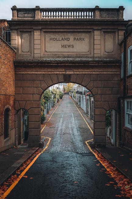 A narrow cobbled street leading through an archway engraved with 'HOLLAND PARK MEWS' on a stone lintel, framed by brick buildings on either side. The street is lined with parked cars, and the overhang