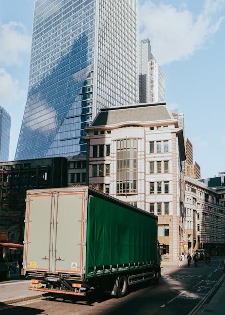 A large green-sided commercial cargo truck is parked on a city street with modern high-rise glass office buildings and older stone structures in the background. The truck is positioned at the curb, wi