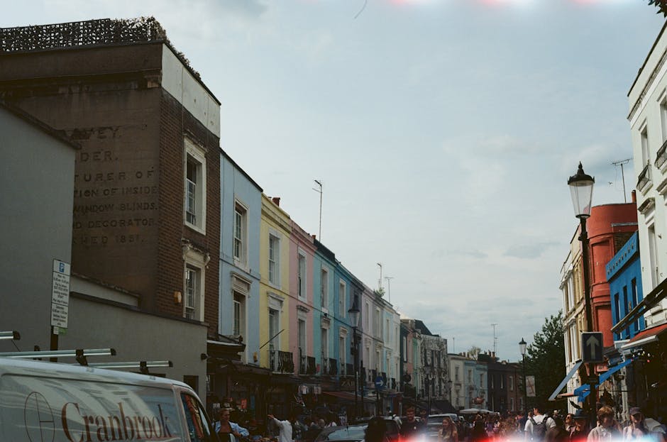 A row of multi-storey Victorian-style terraced houses with brick facades, each painted in different vibrant colors including cream, navy blue, bright green, and pink. The middle house, painted bright green, features large bay windows and a black front door. In front of the houses, there is a tree with bare branches and a tall street lamp, indicating a residential street setting. The image is taken during daylight with clear blue skies. This scene illustrates an urban environment suitable for house removals and relocation services, highlighting the context in which furniture transport and packing and moving activities may take place, consistent with services offered by Man and Van Notting Hill.