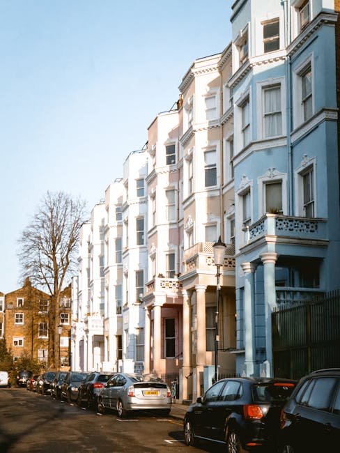 A row of multi-storey residential buildings painted in light pastel colors, including white and pale blue, with bay windows and decorative architectural details, situated along a quiet street in Notting Hill. In front of the buildings, several parked cars occupy the roadside, with some reflecting the sunlight. The street appears calm and clean, with a few leafless trees visible behind the buildings and a clear, blue sky overhead. The scene depicts an exterior view of a typical London neighbourhood, relevant to home relocation and furniture transport services provided by Man and Van Notting Hill, as part of a house removals or packing and moving process.