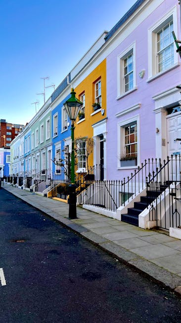 A row of pastel-coloured terraced houses on a residential street in Notting Hill during daylight. The houses feature white-framed sash windows, black wrought iron railings along the front steps, and potted plants on window sills. A traditional black lamp post stands near the pavement, which is made of concrete slabs. The sky is clear and blue, with some television antennas visible on rooftops. The scene depicts a peaceful neighbourhood often associated with home relocation and furniture transport services, with no vehicles or people present but the environment suitable for loading and unloading furniture. This setting may be used to illustrate house removals, packing, or moving logistics in a professional context, highlighting the urban environment where Man and Van Notting Hill offers their removals and relocation services.