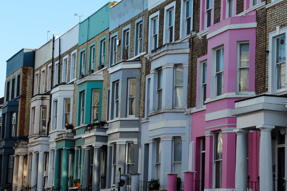 Photograph of a row of Victorian-style terraced houses on a residential street, featuring white and blue facades with ornate bay windows and decorative trim. The house at the forefront has a bold red 